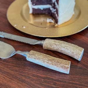 May include: A cake server and knife set with antler-style handles, resting on a dark wood surface. A slice of chocolate cake with white frosting sits on a gold plate in the background. The knife has a serrated blade.