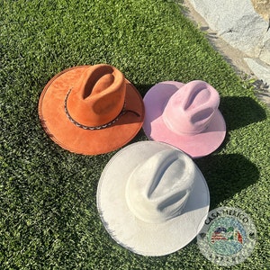 May include: Three felt cowboy hats in different colors: orange, pink, and white. The hats are lying on a green artificial grass surface.