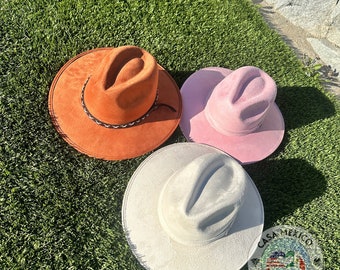 May include: Three felt cowboy hats in different colors: orange, pink, and white. The hats are lying on a green artificial grass surface.
