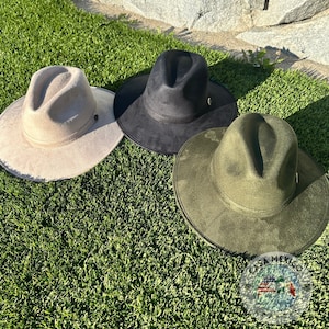 Puede incluir: Tres sombreros fedora de fieltro en diferentes colores: beige, negro y verde oliva. Los sombreros están sobre una superficie de césped artificial verde.