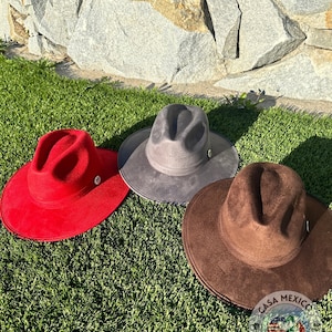 Puede incluir: Tres sombreros fedora de fieltro en diferentes colores: rojo, gris y marrón. Los sombreros están sobre una superficie de césped artificial verde. Los sombreros tienen un pequeño botón plateado en la parte superior.
