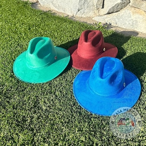 May include: Three felt fedora hats in different colors: teal, burgundy, and royal blue. The hats are sitting on a green grassy surface.