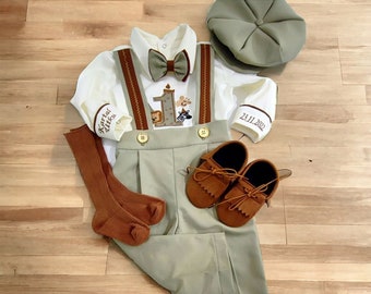 Puede incluir: Un conjunto de bebé para una celebración de primer cumpleaños. El conjunto incluye una camisa blanca con una pajarita marrón, un mono verde con tirantes marrones, un gorro verde, calcetines marrones y mocasines marrones. La camisa tiene un número uno bordado y la fecha 23.11.2020. El texto "Karsten Otto" también está bordado en la camisa.