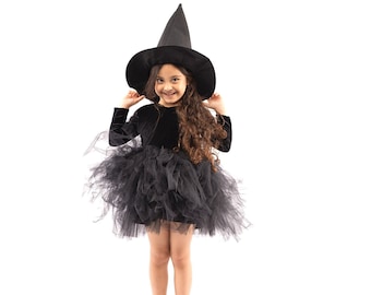 Puede incluir: Una niña vestida de bruja para Halloween. Lleva un vestido de terciopelo negro con una falda de tul negro y un sombrero de bruja negro. Está sonriendo y mirando a la cámara.
