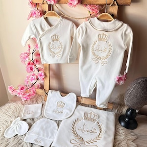 May include: A white baby outfit set with a crown and Arabic text embroidery. The set includes a long-sleeved onesie, a wrap-style shirt, a bib, a hat, a pair of mittens, and a blanket.