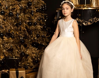 May include: A young girl wearing a white tulle dress with a satin bodice and a pearl headband stands in front of a gold Christmas tree. She is holding the skirt of her dress with one hand.