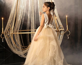 May include: A young girl wearing a white and beige tulle dress with a satin bodice and a bow at the back. She is standing in front of a large, ornate chandelier with crystal beads.
