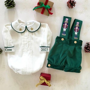 Puede incluir: Una camisa blanca de botones con ribetes verdes y estrellas doradas en el cuello. La camisa tiene el texto "Crew" en un puño y "2023" en el otro. Un mono de terciopelo verde con un diseño de cascanueces en las correas. El mono tiene botones dorados y está doblado por la mitad.
