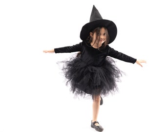 Puede incluir: Una niña vestida de bruja para Halloween. Lleva un vestido de terciopelo negro con una falda de tul negro y un sombrero de bruja negro. Está sonriendo y mirando hacia un lado.