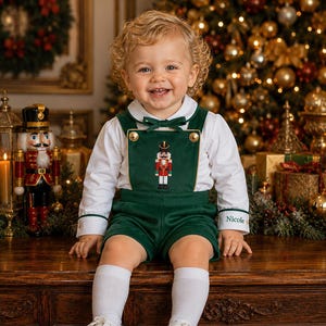 May include: A dark green overall short set with a white long-sleeve collared shirt. The overalls feature a gold-trimmed bib with a nutcracker embroidery, gold buttons, and a small bow at the neckline. The name Nicole is embroidered on the cuff.