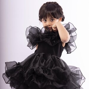 K&ouml;nnte beinhalten: Ein junges M&auml;dchen tr&auml;gt ein schwarzes T&uuml;llkleid mit mehreren R&uuml;schen und einem gro&szlig;en R&uuml;schenkragen. Das Kleid ist &auml;rmellos und hat einen taillierten Oberteil.