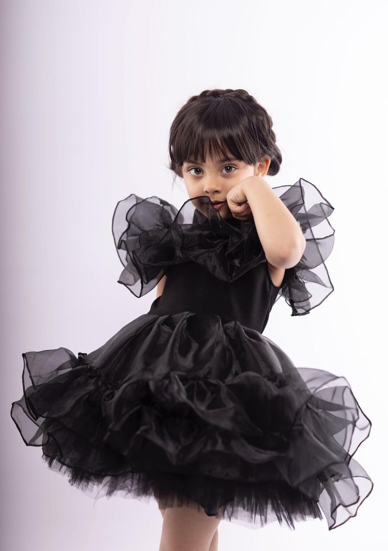 K&ouml;nnte beinhalten: Ein junges M&auml;dchen tr&auml;gt ein schwarzes T&uuml;llkleid mit mehreren R&uuml;schen und einem gro&szlig;en R&uuml;schenkragen. Das Kleid ist &auml;rmellos und hat einen taillierten Oberteil.
