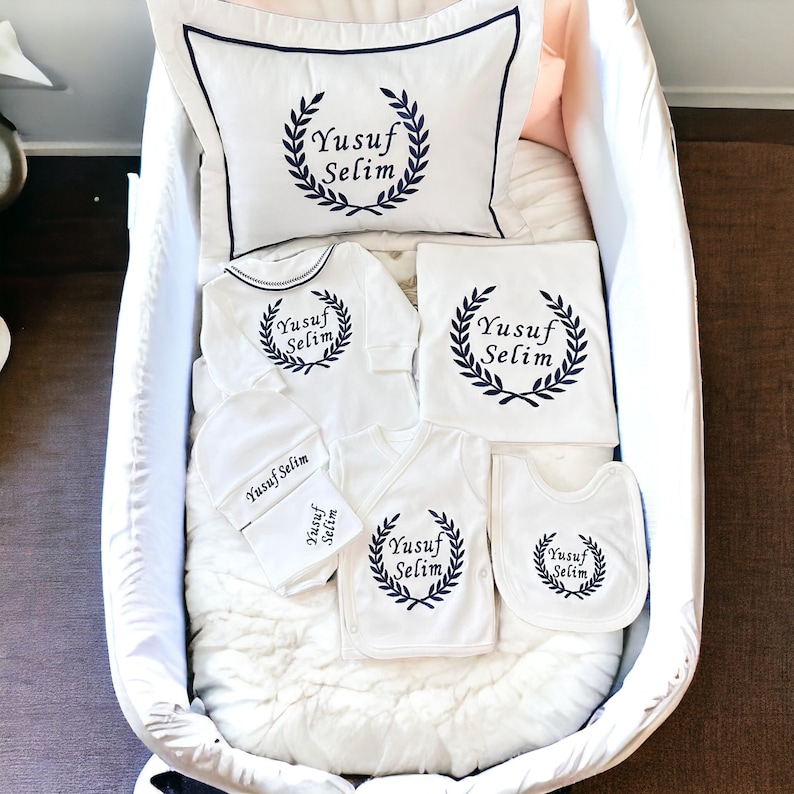 May include: A white baby gift set with a white pillow, a white onesie, a white bib, a white hat, and a white blanket. All items feature a navy blue wreath with the text "Yusuf Selim" in a cursive font.
