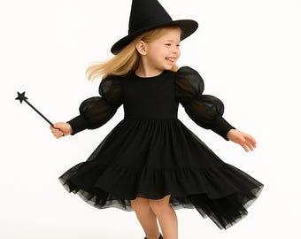 Puede incluir: Un disfraz de bruja negro para niña, que incluye un vestido con mangas transparentes y una falda escalonada. El atuendo incluye un sombrero puntiagudo y una varita con estrella. La niña lleva zapatos negros.