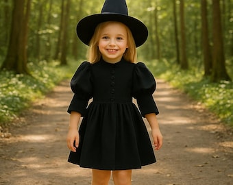 May include: A young child wearing a black witch costume, including a dress, hat, and shoes, stands on a path in a forest. The dress has a high collar and button details. The hat is a classic pointed design.
