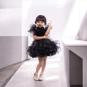 K&ouml;nnte beinhalten: Ein junges M&auml;dchen tr&auml;gt ein schwarzes T&uuml;llkleid mit R&uuml;schen. Sie steht in einem wei&szlig;en Raum und l&auml;chelt in die Kamera.