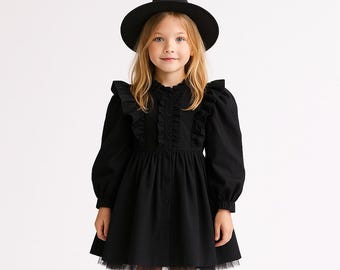 Puede incluir: Una niña pequeña con un disfraz de bruja negro, que incluye un vestido con volantes y un dobladillo de tul, y un sombrero puntiagudo. El vestido tiene mangas largas y botones en la parte delantera. El fondo es blanco liso.