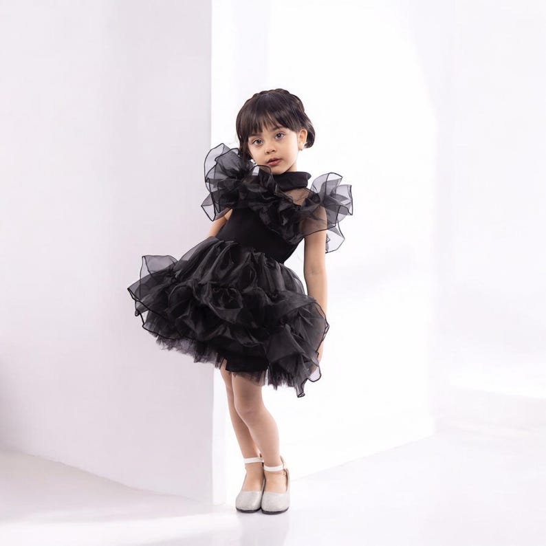 K&ouml;nnte beinhalten: Ein junges M&auml;dchen tr&auml;gt ein schwarzes T&uuml;llkleid mit mehreren R&uuml;schenlagen. Das Kleid hat einen hohen Ausschnitt und kurze &Auml;rmel. Das M&auml;dchen steht in einem wei&szlig;en Raum und schaut in die Kamera.
