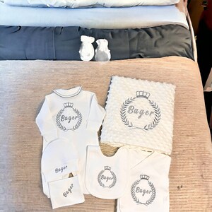 May include: A white baby outfit set with a long-sleeved onesie, a bib, a hat, a burp cloth, and a blanket. All items feature a gray embroidered crown and the name "Bager" in a wreath.