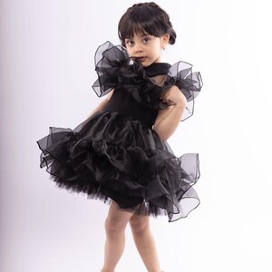 K&ouml;nnte beinhalten: Ein junges M&auml;dchen tr&auml;gt ein schwarzes T&uuml;llkleid mit R&uuml;schen und silbernen Schuhen. Das Kleid hat einen hohen Ausschnitt und ist &auml;rmellos.