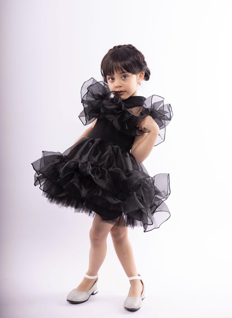 K&ouml;nnte beinhalten: Ein junges M&auml;dchen tr&auml;gt ein schwarzes T&uuml;llkleid mit R&uuml;schen und silbernen Schuhen. Das Kleid hat einen hohen Ausschnitt und ist &auml;rmellos.