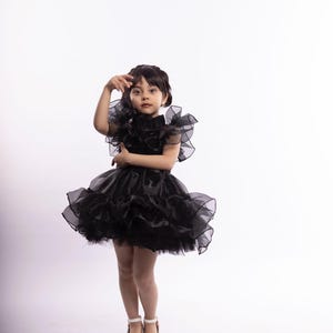 K&ouml;nnte beinhalten: Ein junges M&auml;dchen tr&auml;gt ein schwarzes T&uuml;llkleid mit mehreren Lagen und R&uuml;schen. Sie steht vor einem wei&szlig;en Hintergrund und hat die Hand am Kopf.
