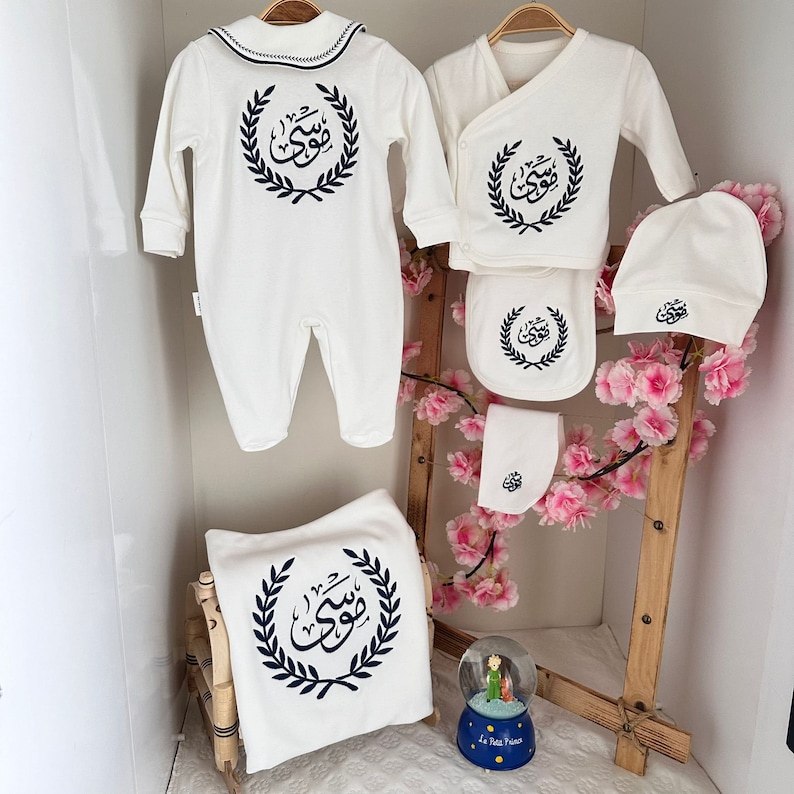 May include: A white baby outfit set with a navy blue embroidered design featuring Arabic calligraphy. The set includes a onesie, a hat, a bib, and a burp cloth. The design features a wreath with the Arabic text "Moudy" in the center.
