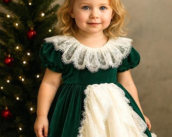 May include: A young child wearing a green velvet dress with a white lace collar and skirt detail. The dress has short sleeves and a ruffled hem. A Christmas tree with red ornaments is in the background.