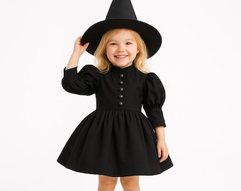 Puede incluir: Un niño con un disfraz de bruja negro, que incluye un vestido con cuello alto y detalles de botones, y un sombrero puntiagudo. El vestido tiene mangas largas y una falda acampanada. El niño sonríe y mira a la cámara.