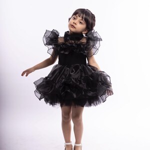 K&ouml;nnte beinhalten: Ein junges M&auml;dchen tr&auml;gt ein schwarzes T&uuml;llkleid mit R&uuml;schen und einem hohen Kragen. Sie steht auf einem wei&szlig;en Hintergrund und schaut in die Kamera.