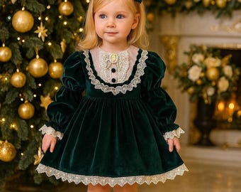 May include: A little girl in a dark green velvet dress with white lace trim and a matching green bow. The dress has long sleeves and a high neckline. She is wearing black Mary Jane shoes. A Christmas tree with gold ornaments is in the background.