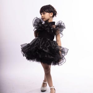 K&ouml;nnte beinhalten: Ein junges M&auml;dchen tr&auml;gt ein schwarzes T&uuml;llkleid mit mehreren R&uuml;schenlagen. Das Kleid hat einen hohen Kragen und kurze &Auml;rmel. Sie tr&auml;gt wei&szlig;e Schuhe mit Kn&ouml;chelriemen.