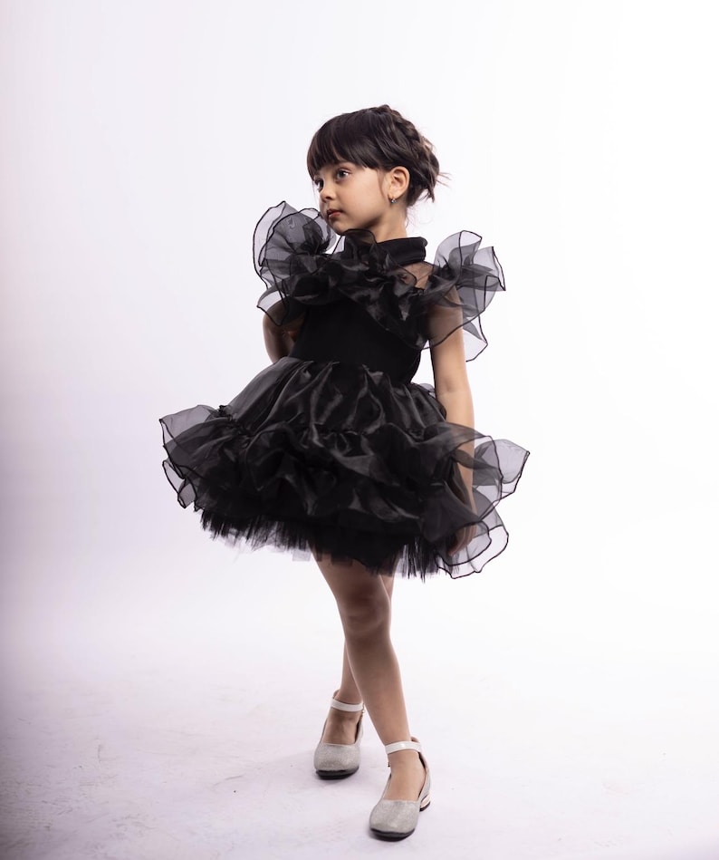 K&ouml;nnte beinhalten: Ein junges M&auml;dchen tr&auml;gt ein schwarzes T&uuml;llkleid mit mehreren R&uuml;schenlagen. Das Kleid hat einen hohen Kragen und kurze &Auml;rmel. Sie tr&auml;gt wei&szlig;e Schuhe mit Kn&ouml;chelriemen.