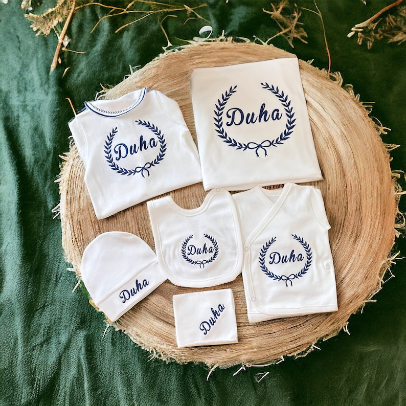 May include: A set of white baby clothes with the name "Duha" embroidered in blue on each piece. The set includes a onesie, a bib, a hat, a burp cloth, and a swaddle blanket.