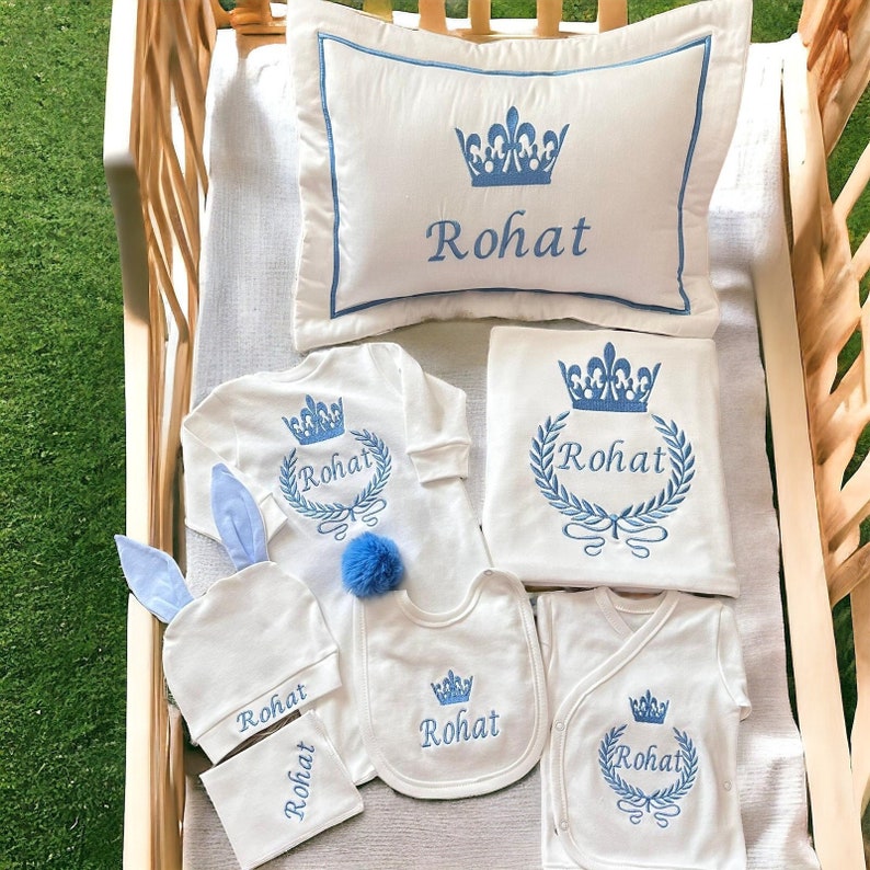 May include: A baby gift set with a blue and white color scheme. The set includes a pillow, blanket, bib, onesie, hat, and a small towel. All items feature a crown and the name "Rohat" embroidered in blue.