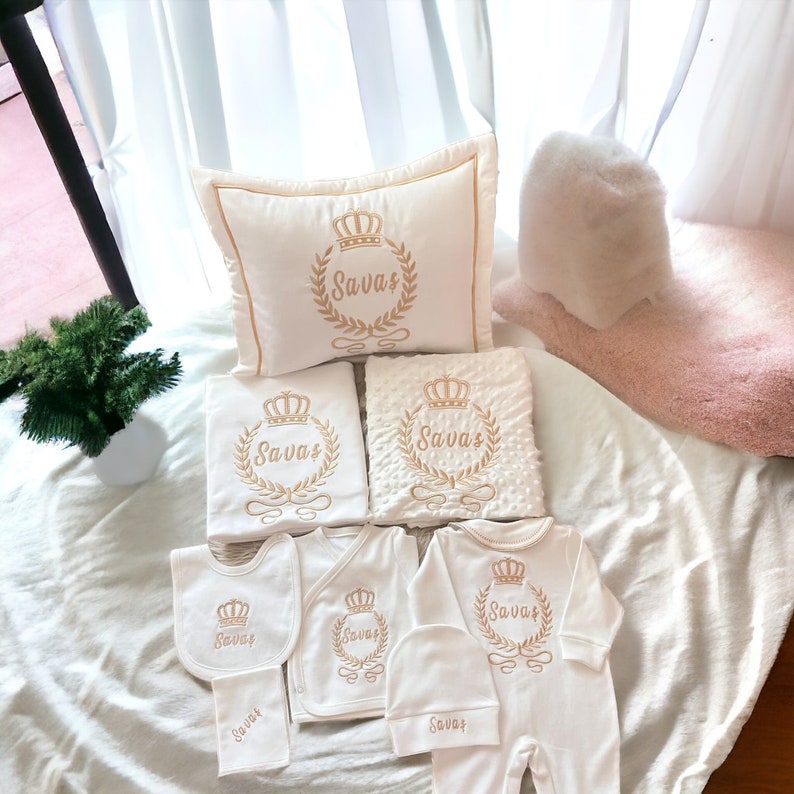 May include: A white baby gift set with gold embroidery featuring the name "Savvas" and a crown design. The set includes a pillow, blanket, bib, hat, and two outfits.