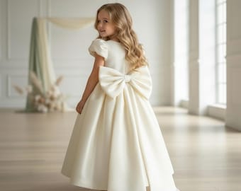 Op de afbeelding: Een jong meisje draagt een ivoorkleurige satijnen jurk met een grote strik op de rug. De jurk heeft korte mouwen en een zwierige rok met een sleep. Het meisje heeft lang, golvend blond haar en kijkt over haar schouder.