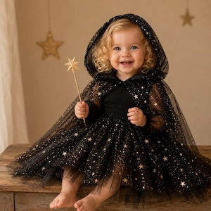 May include: A black tulle dress featuring a hood and a skirt adorned with gold glitter stars. The garment includes long sheer sleeves and a matching black bodice. A gold star-shaped wand is held in one hand, completing the celestial-themed outfit.