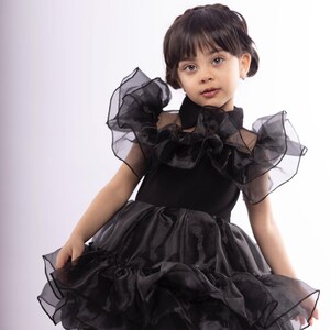 K&ouml;nnte beinhalten: Ein junges M&auml;dchen tr&auml;gt ein schwarzes T&uuml;llkleid mit mehreren R&uuml;schenlagen. Das Kleid hat einen hohen Kragen und kurze &Auml;rmel. Das M&auml;dchen schaut mit ernster Miene in die Kamera.
