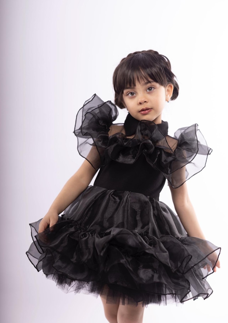 K&ouml;nnte beinhalten: Ein junges M&auml;dchen tr&auml;gt ein schwarzes T&uuml;llkleid mit mehreren R&uuml;schenlagen. Das Kleid hat einen hohen Kragen und kurze &Auml;rmel. Das M&auml;dchen schaut mit ernster Miene in die Kamera.
