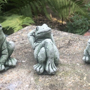 May include: Three stone frog ornaments in different poses. One sits upright, another rests a hand on its head, and the third sits with its legs tucked. The statues are a muted green colour and appear weathered, suggesting a garden theme.