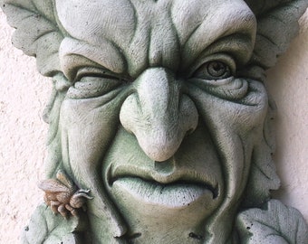 Op de afbeelding: Een gedetailleerde, verweerde groene man gezicht sculptuur met blad-achtige kenmerken. Het gezicht heeft een knipogend oog, een prominente neus en een gedetailleerde mond. Een kleine bij en lieveheersbeestje zijn zichtbaar op de bladeren. Tuindecoratie.
