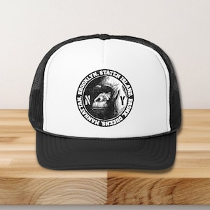 Puede incluir: Gorra de camionero en blanco y negro con un panel frontal blanco que presenta un círculo con el texto "MANHATTAN, QUEENS, BROOKLYN, STATEN ISLAND, BRONX" y una cabeza de gorila dentro del círculo. El texto "NY" está dentro del círculo sobre la cabeza del gorila. La gorra tiene un panel trasero de malla negra.