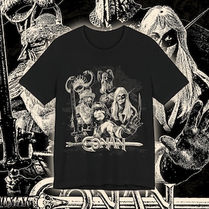 Könnte beinhalten: Schwarzes T-Shirt mit einer Schwarz-Weiß-Grafik von Charakteren aus Conan der Barbar. Das Wort "CONAN" wird in einer stilisierten Schriftart am unteren Rand der Grafik angezeigt. Das T-Shirt ist kurzärmelig.