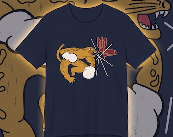 May include: Navy blue t-shirt featuring a cartoon jaguar with white boxing gloves and a red cactus. The jaguar is in a fighting stance, with a white burst emanating from its mouth. The background includes a larger jaguar illustration.