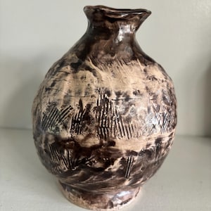 May include: A round ceramic vase with a narrow neck and a flared rim. The surface features a textured, mottled pattern in shades of dark brown and cream, with visible vertical etched lines and a glossy finish covering the entire exterior body.