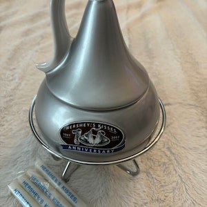 May include: Silver Hershey's Kisses chocolate fondue pot with a stand and four dipping forks. The pot has a 100th anniversary logo with the years 1907 and 2007.