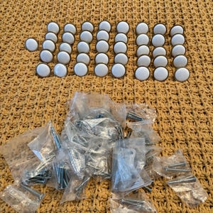 May include: A set of 30 white ceramic cabinet knobs with gold trim and a bag of screws. The knobs are arranged in rows on a yellow knitted surface.