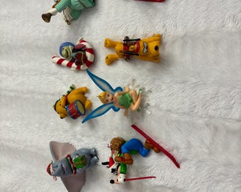 Può includere: Una collezione di decorazioni natalizie con vari personaggi Disney. Gli ornamenti includono personaggi come Peter Pan su una slitta, Dumbo, Winnie the Pooh, Trilli e Paperino, tutti in abiti festivi. Gli ornamenti sono in plastica e colorati.