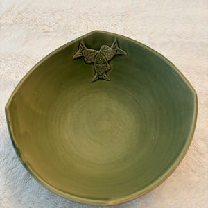 May include: A green ceramic bowl with a unique, angular shape. The bowl features a raised relief design of three fish in the center. The bowl's interior has a smooth, matte finish, and the exterior has a slightly textured appearance.